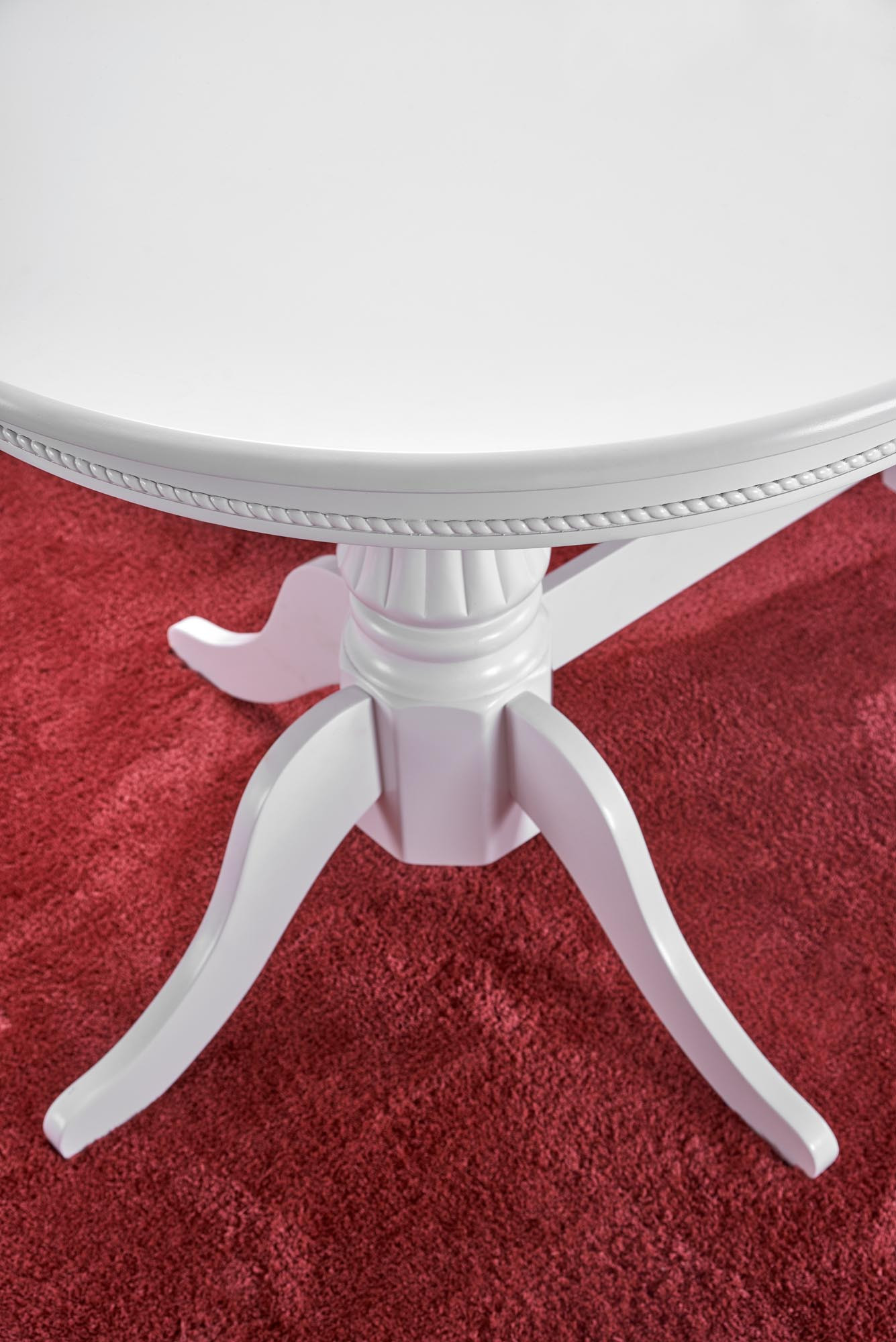 Zbliżenie na detal białego rozkładanego stołu Jonah z ozdobnym wykończeniem na czerwonym dywanie.
